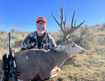 A rifle hunter with his mule deer trophy smiles proudly on a beautiful fall day.